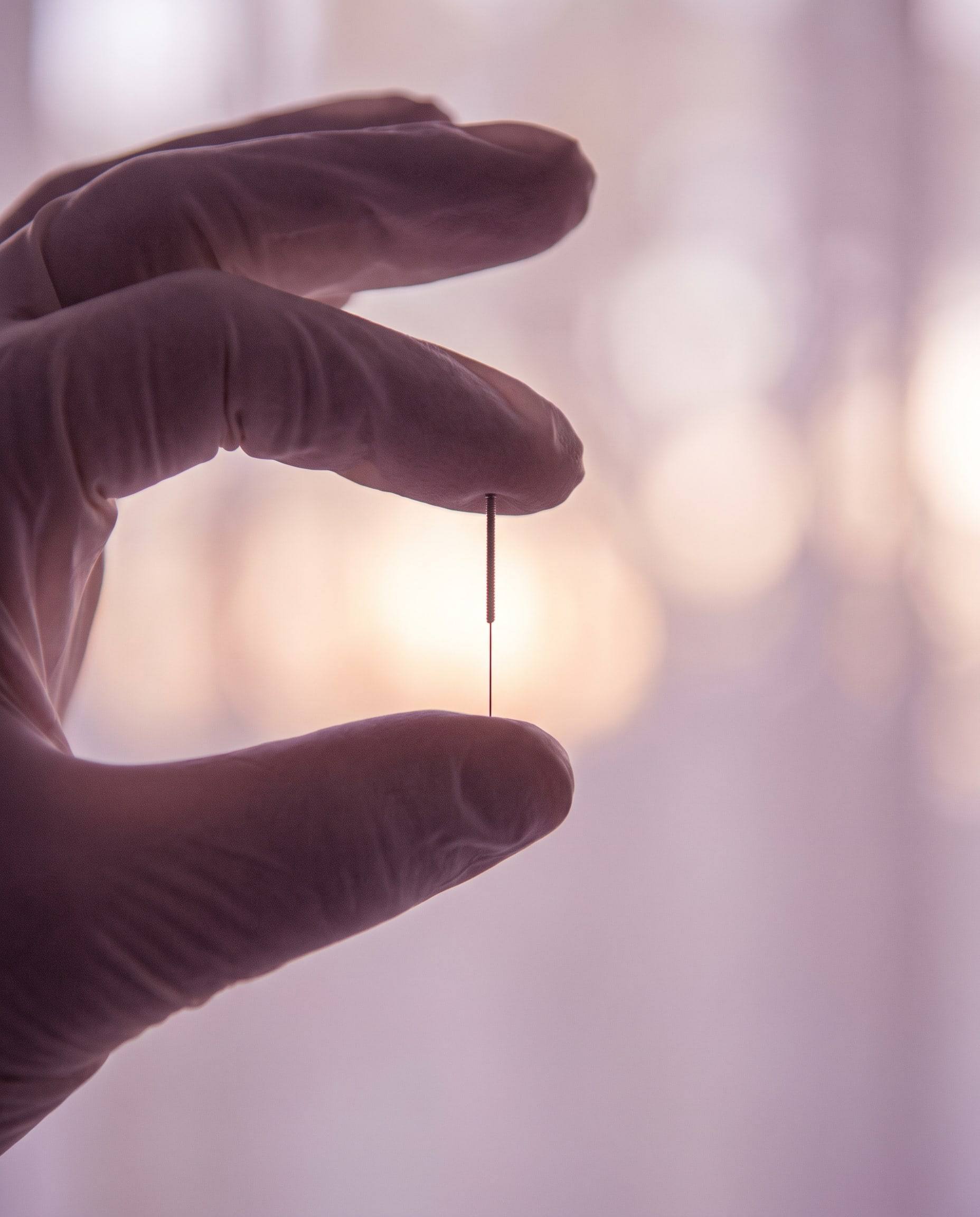 A gloved hand holding a single fine acupuncture needle against soft light
